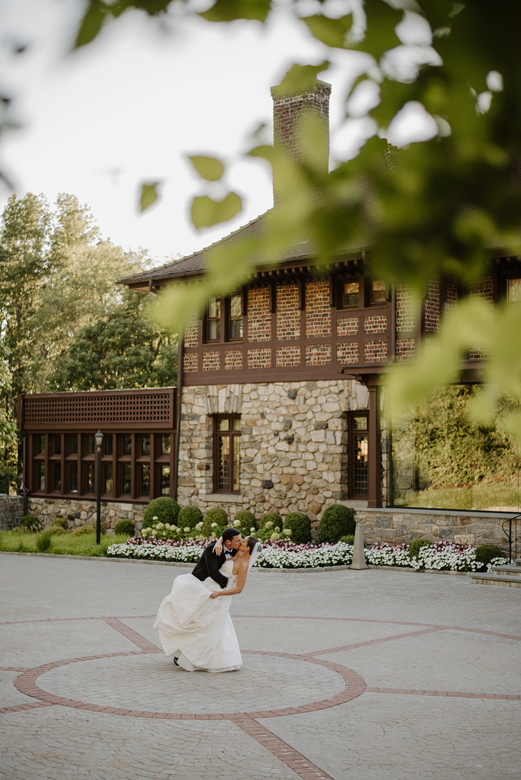 Groom dipping and kissing the bride in the mansion courtyard, framed by soft green leaves in the foreground.